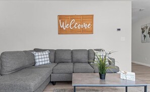 Ein modernes Wohnzimmer mit einem grauen Ecksofa, einem Holz-Willkommen-Schild, einem kleinen Tisch mit einer Pflanze und einer Weltkarte an der Wand.