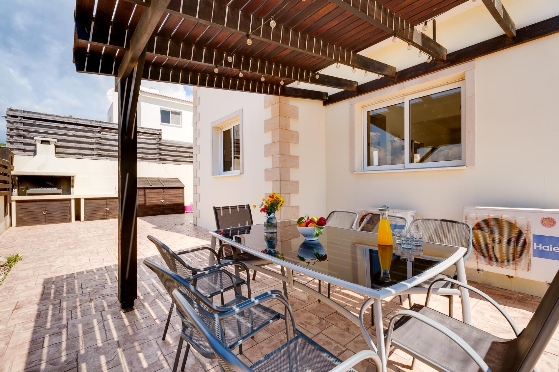 Um pátio externo com mesa de vidro, cadeiras de metal, churrasqueira e vista para um edifício residencial.