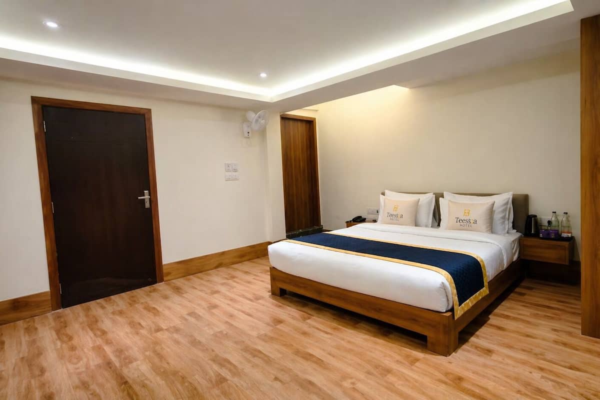 A hotel room with a wooden floor, a bed with white bedding and a blue accent, a nightstand with a phone and a bottle, and a door with a wooden frame.