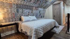 Een slaapkamer met een bed, een nachtkastje, een muur met kentekens, een deur en een bureau met een lamp.