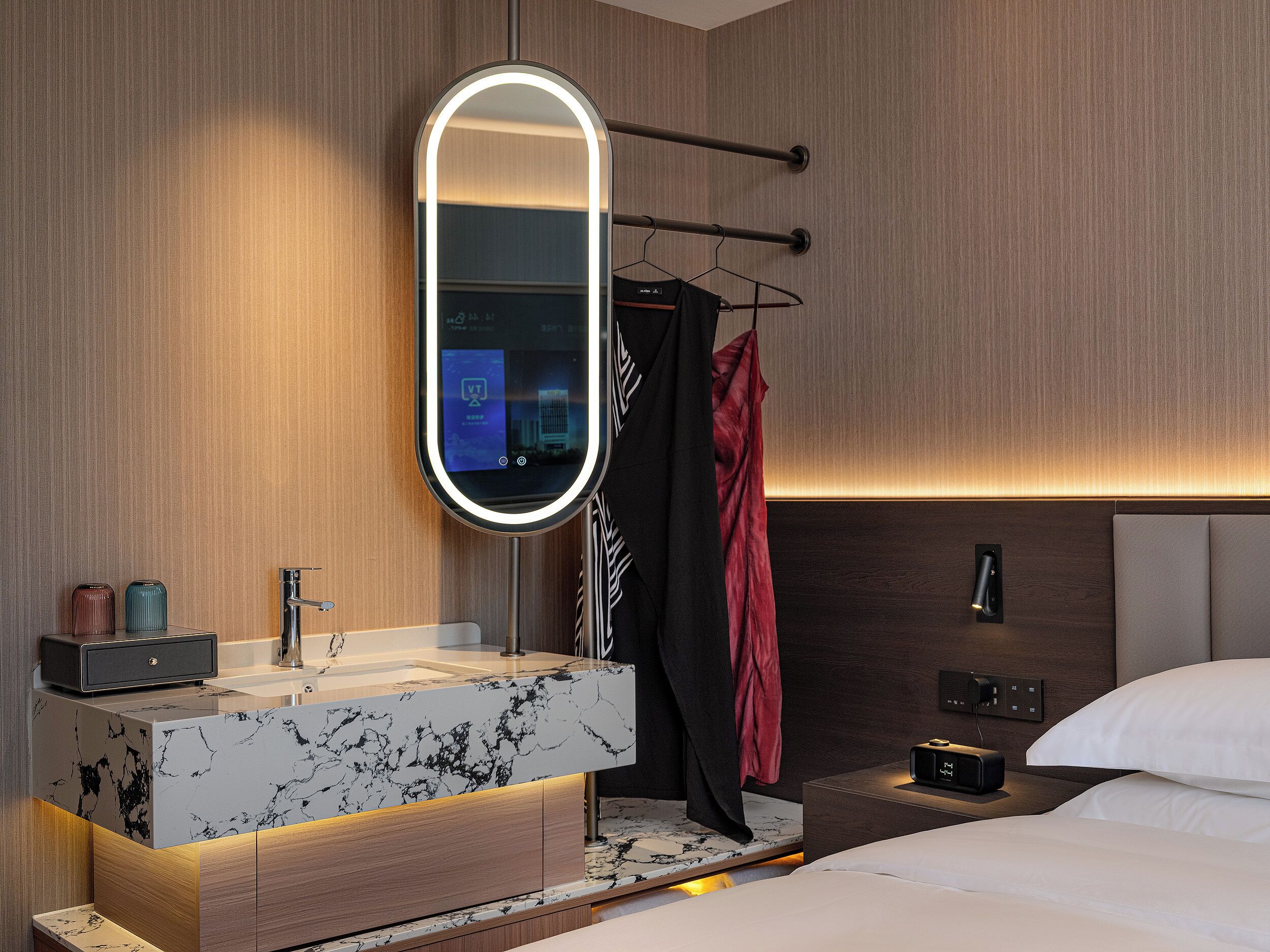 A modern bathroom with a marble vanity, a large mirror, a towel rack, and a bedside table with a lamp.