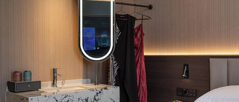 A modern bathroom with a marble vanity, a large mirror, a towel rack, and a bedside table with a lamp.