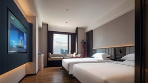 Habitación de hotel con un ventanal grande, dos camas, un escritorio con una computadora portátil, una silla y una televisión mostrando un paisaje urbano.