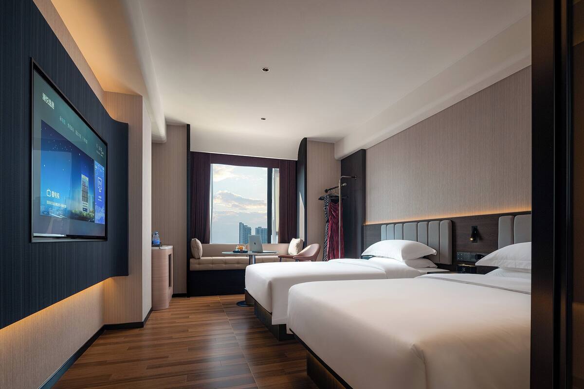 A hotel room with a large window, two beds, a desk with a laptop, a chair, and a TV screen displaying a cityscape.