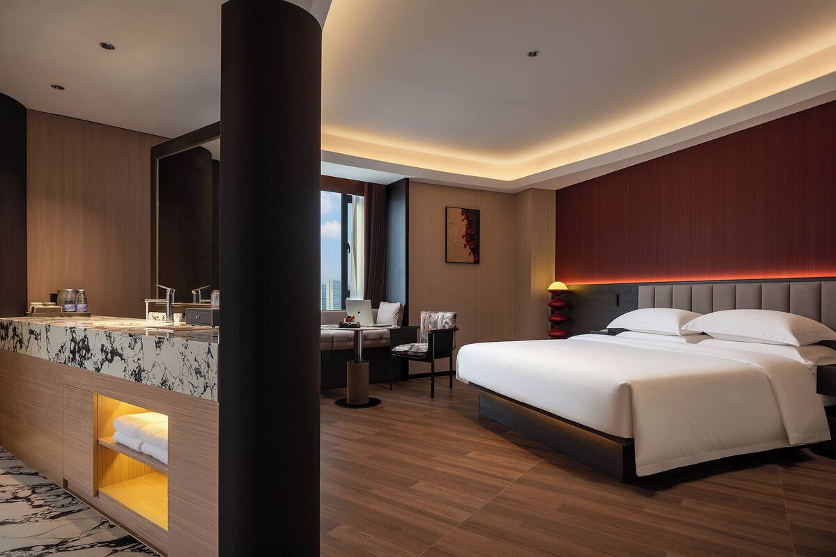 A modern hotel room with a bed, desk, and seating area, featuring a city view.