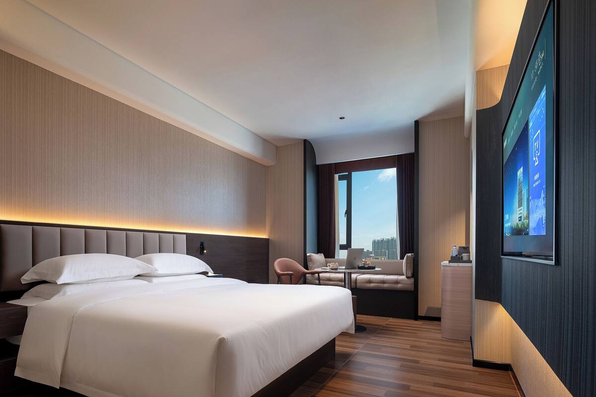 A modern hotel room with a large bed, a seating area with a laptop, and a large window offering a city view.