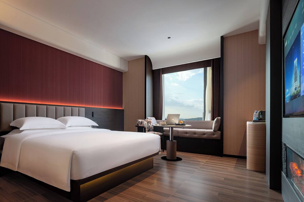 A modern hotel room with a large bed, a desk with a laptop, a sofa, and a large window offering a city view.
