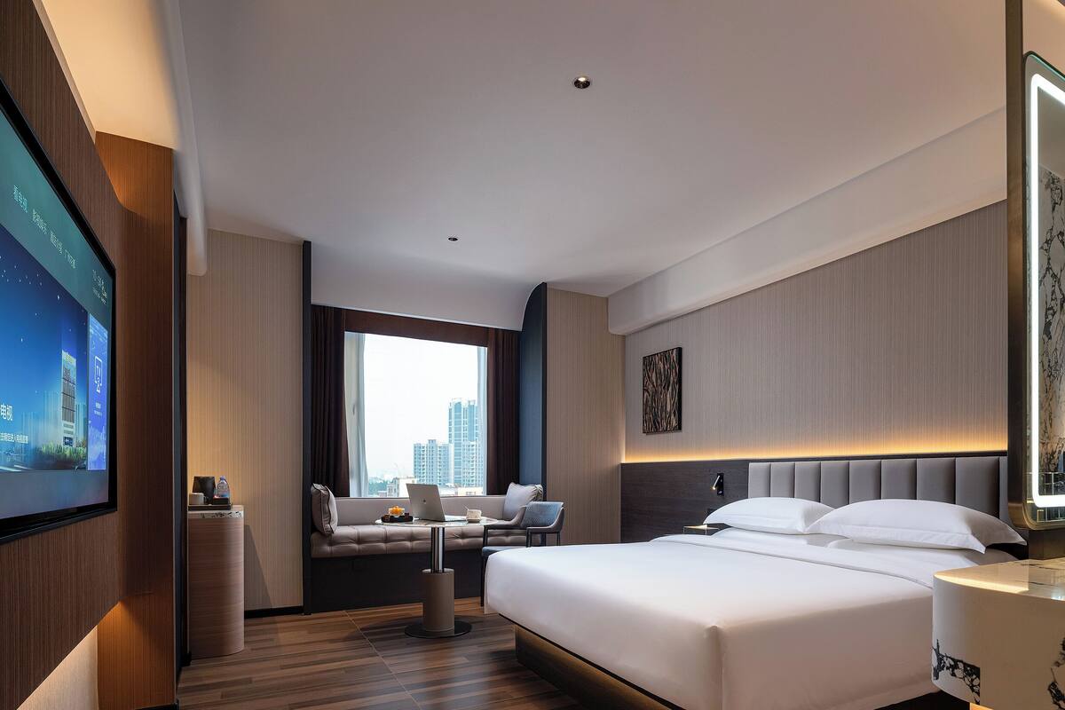 A modern hotel room with a large bed, a seating area with a sofa and armchairs, a television, and a city view.