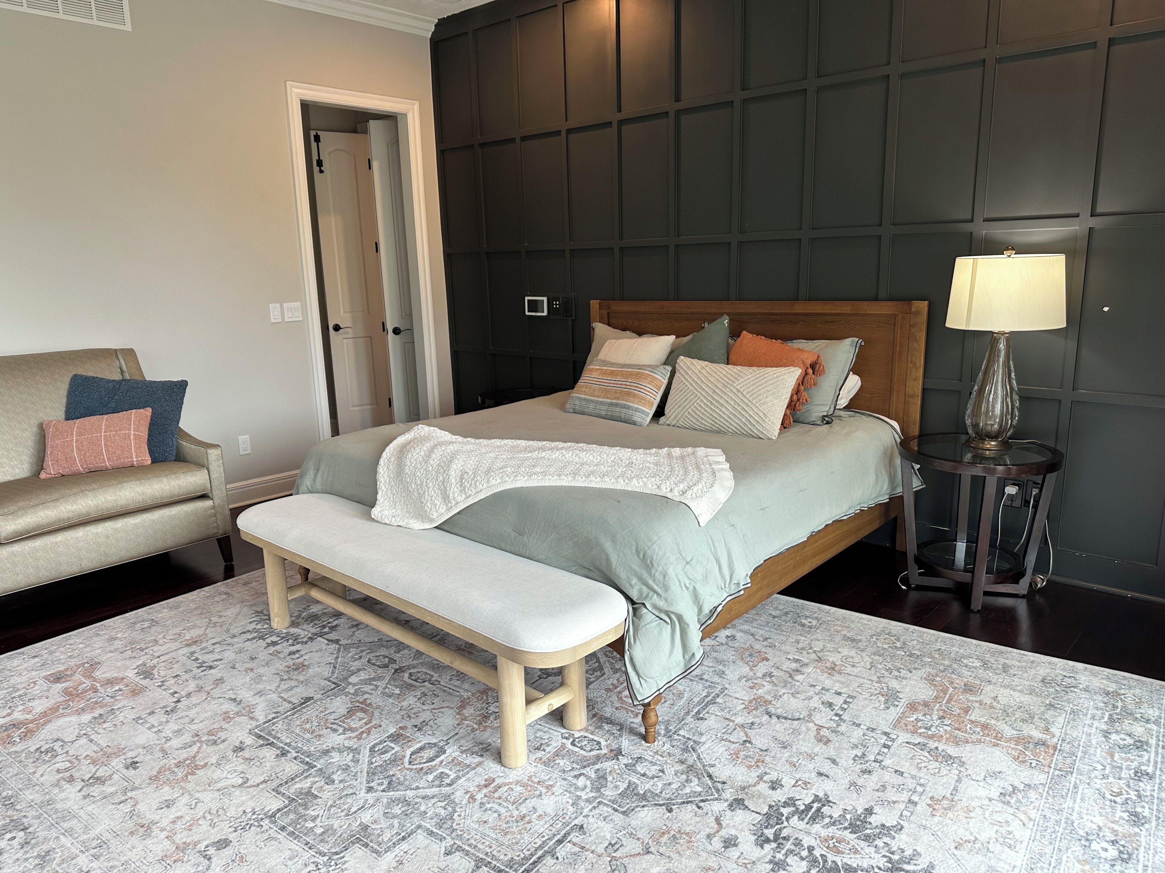 A bedroom with a bed, a bench, a chair, a bedside table with a lamp, and a patterned rug.