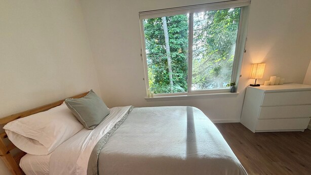Quarto com cama, mesa de cabeceira, janela com vista para árvores e luminária.