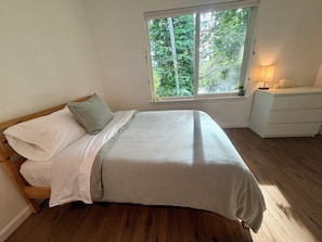 Ein Schlafzimmer mit einem Bett, einem Nachttisch, einem Fenster mit Blick auf Bäume und einer Lampe.