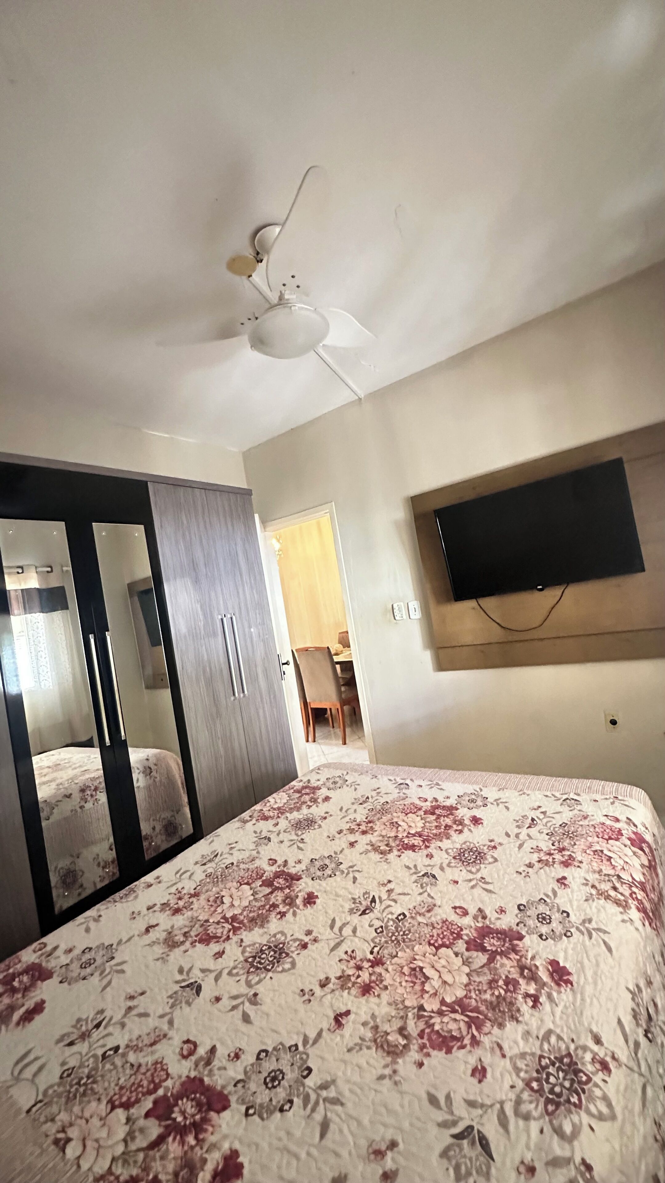 Un dormitorio con cama, armario, ventilador de techo, televisor montado en la pared y una puerta que da a otra habitación.