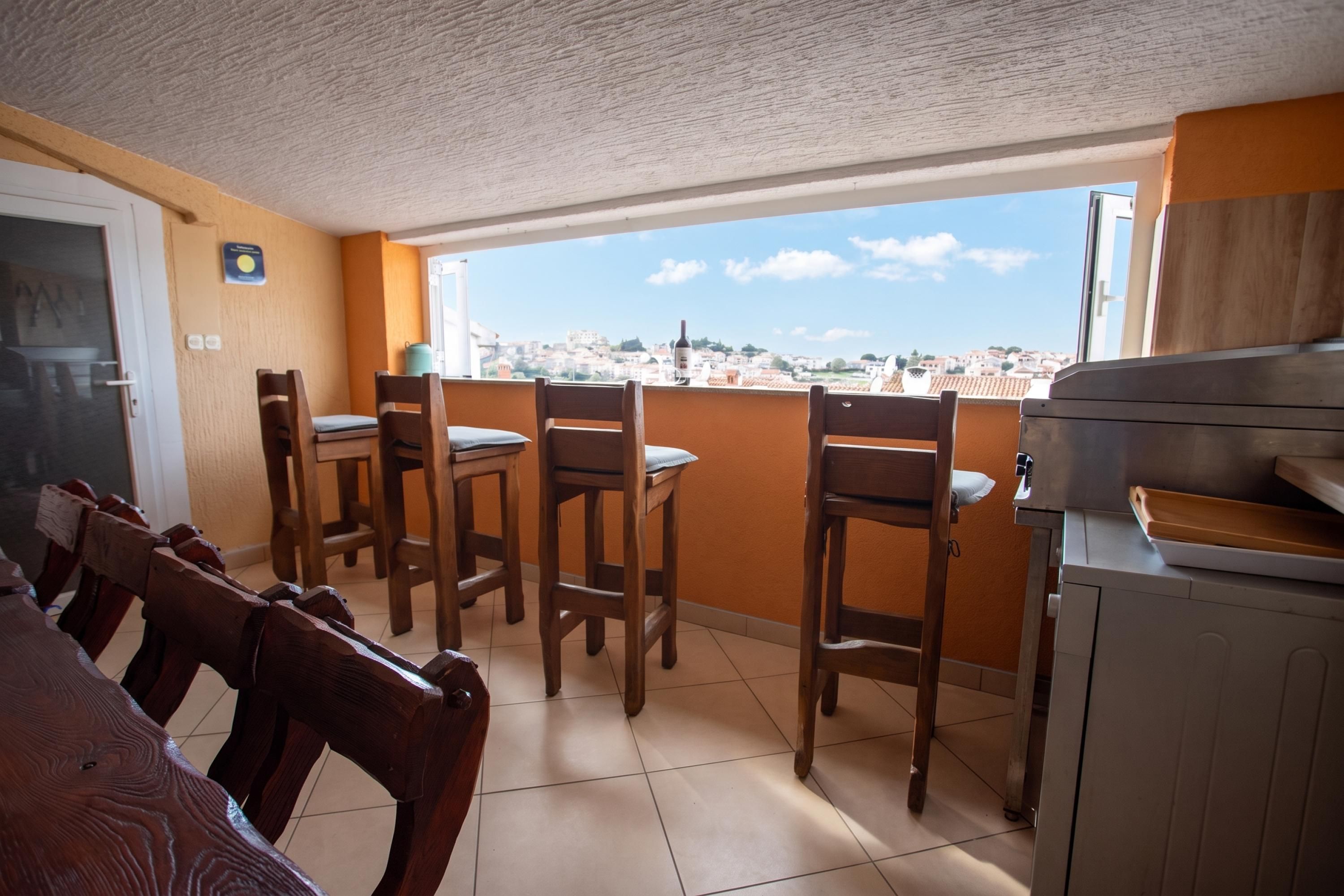 A dining area with wooden chairs and a table, a kitchen with a stove, and a view of a cityscape through a window.