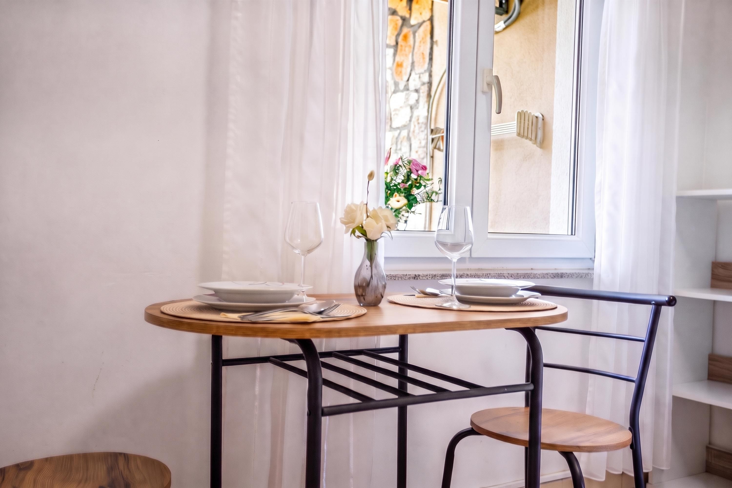 A dining table with plates, glasses, and a vase with flowers, set against a window with curtains.
