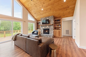 A spacious living room with a stone fireplace, wooden ceiling, and large windows.