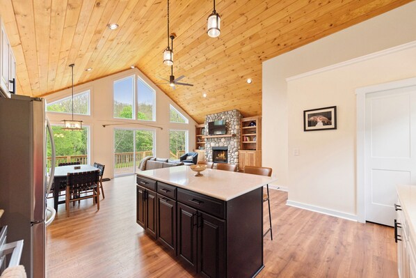 A spacious kitchen with a large island, wooden ceiling, and a stone fireplace.