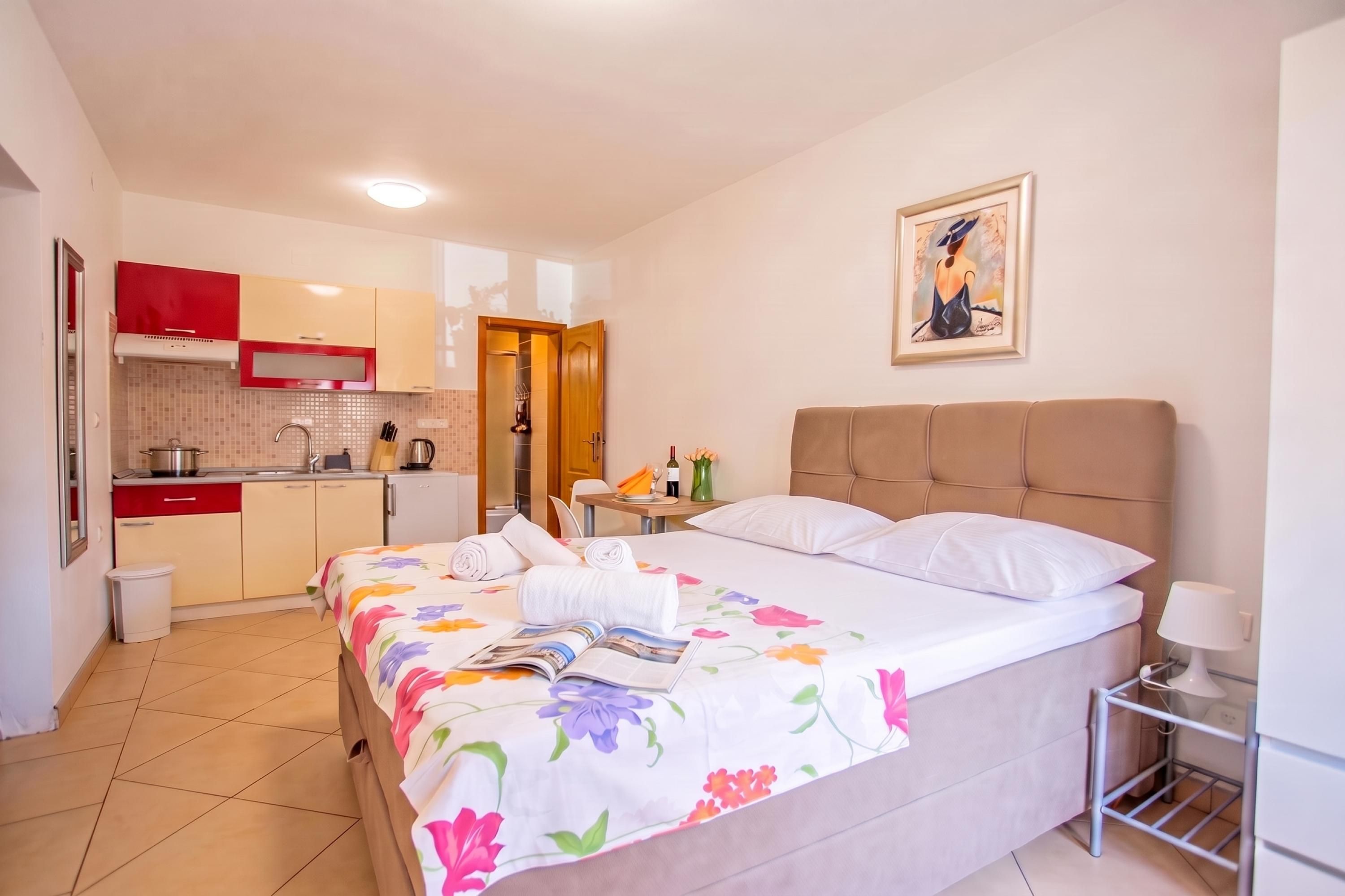 A hotel room with a bed, a bedside table with a lamp, a small kitchen area, and a door leading to a bathroom.