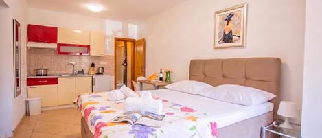 A hotel room with a bed, a bedside table with a lamp, a small kitchen area, and a door leading to a bathroom.
