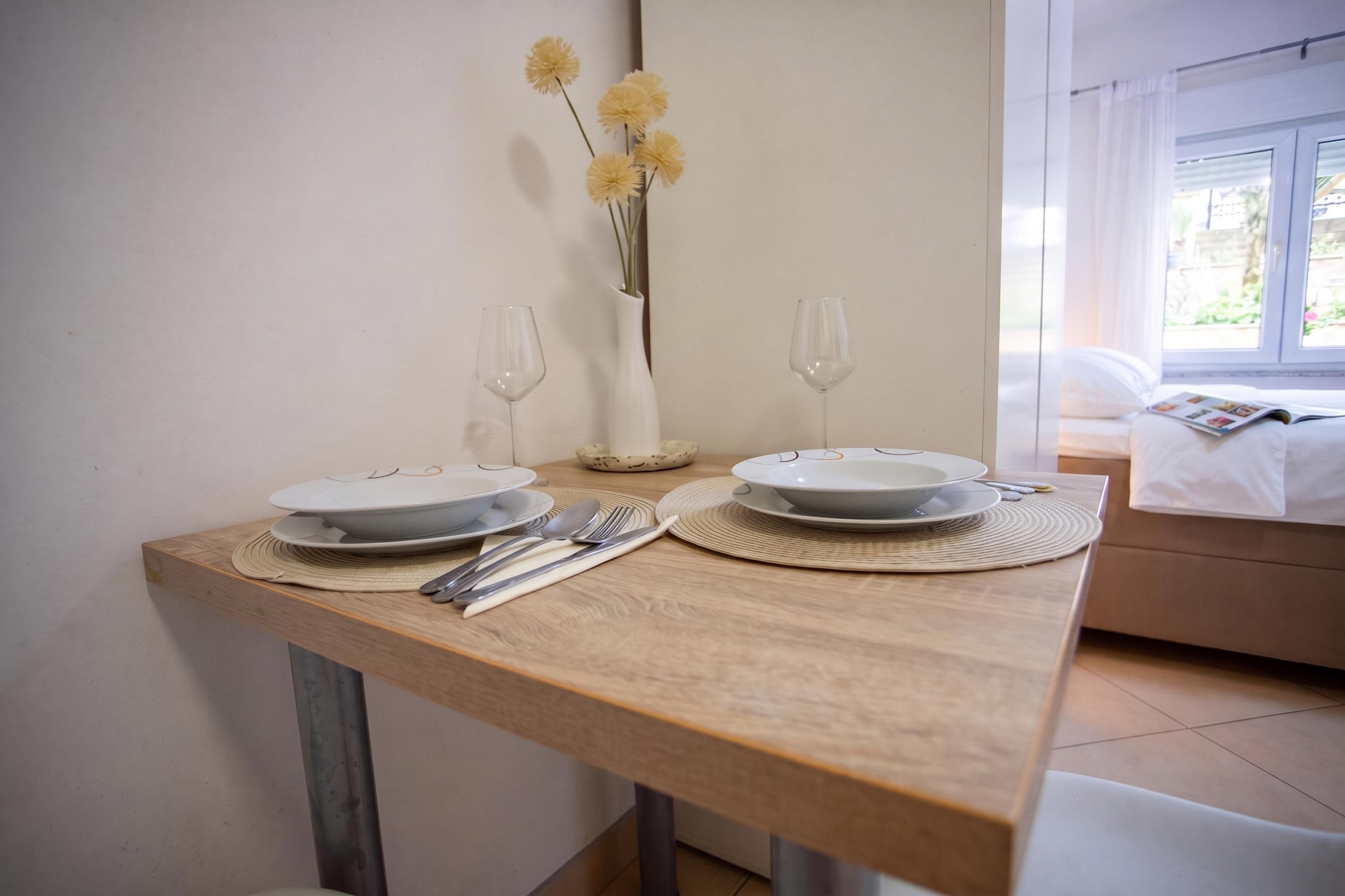 Una mesa de comedor dispuesta con platos, cubiertos y dos copas de vino, con un florero en la pared de fondo.
