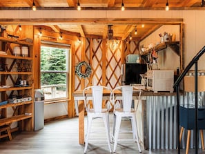 A rustic kitchen with wooden beams, a large window, and a dining area with white chairs. - Starlight Yurt- A Romantic Stay w/ Hot Tub Views (Fancy Gap)