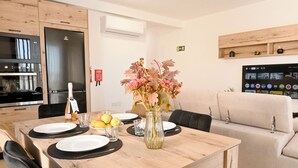 A dining area with a wooden table, chairs, a vase with flowers, and a kitchen in the background. - Viviani Court 2bd Modern Apt Xewkija Gozo, XEWKIJA Gozo, Malta (XEWKIJA Gozo)