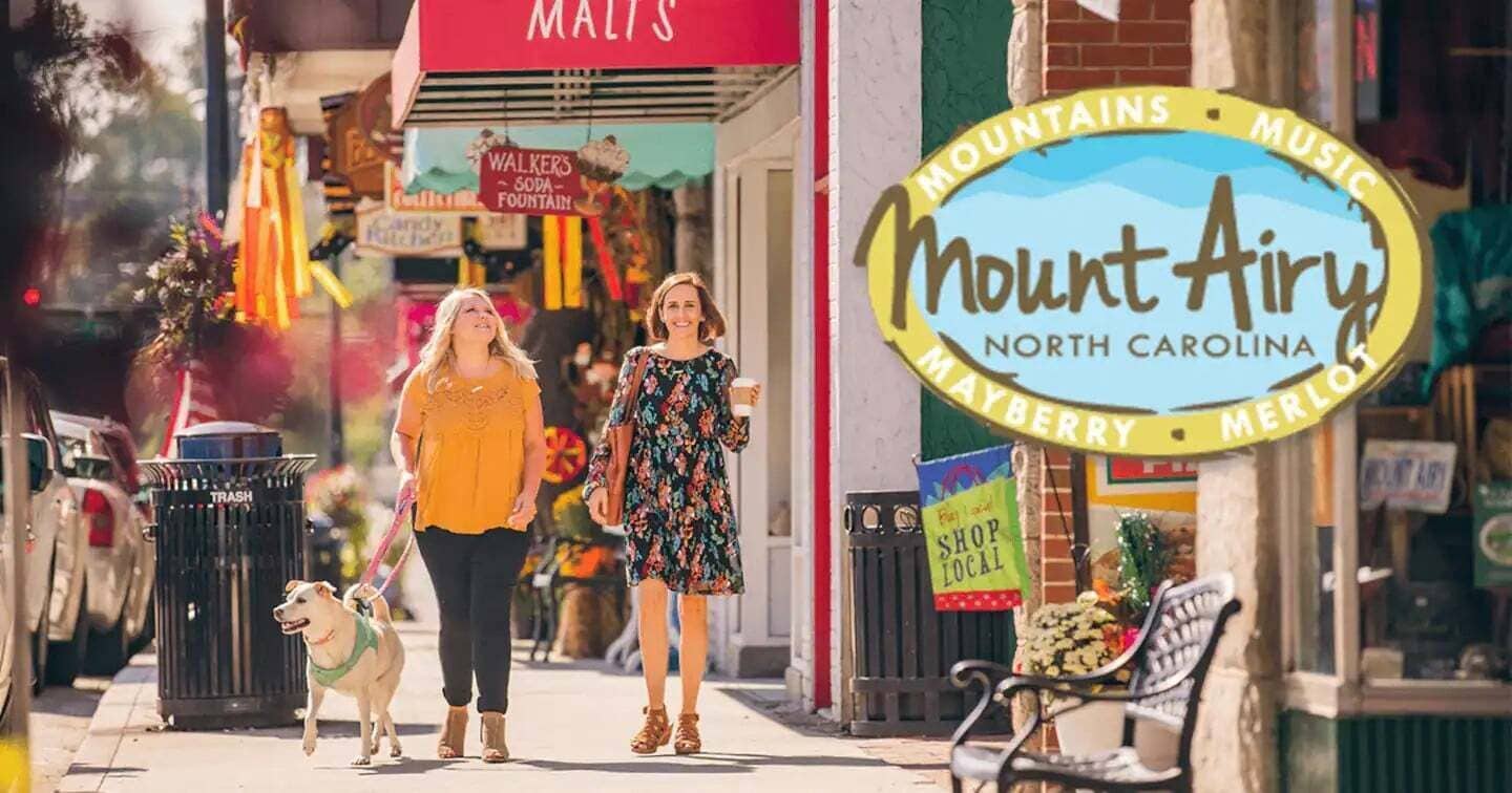 Una scena di strada con un cartello che indica 'Mount Airy North Carolina' e uno striscione con scritto 'Acquista dai commercianti locali'.