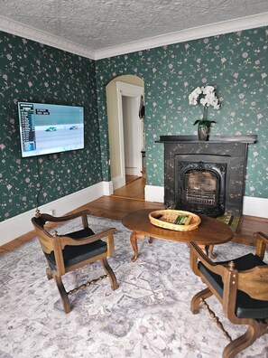 A room with a fireplace, floral wallpaper, a television displaying a racing game, and a dining area with a table and chairs.