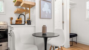 Une cuisine moderne avec des meubles blancs, une table ronde noire, deux chaises blanches, des étagères en bois au-dessus de l’évier et une porte donnant sur une autre pièce.