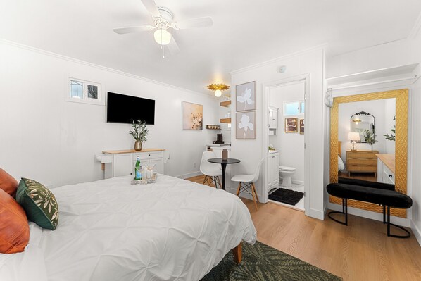 Un dormitorio con cama, escritorio, silla, televisor y baño con espejo y retrete.