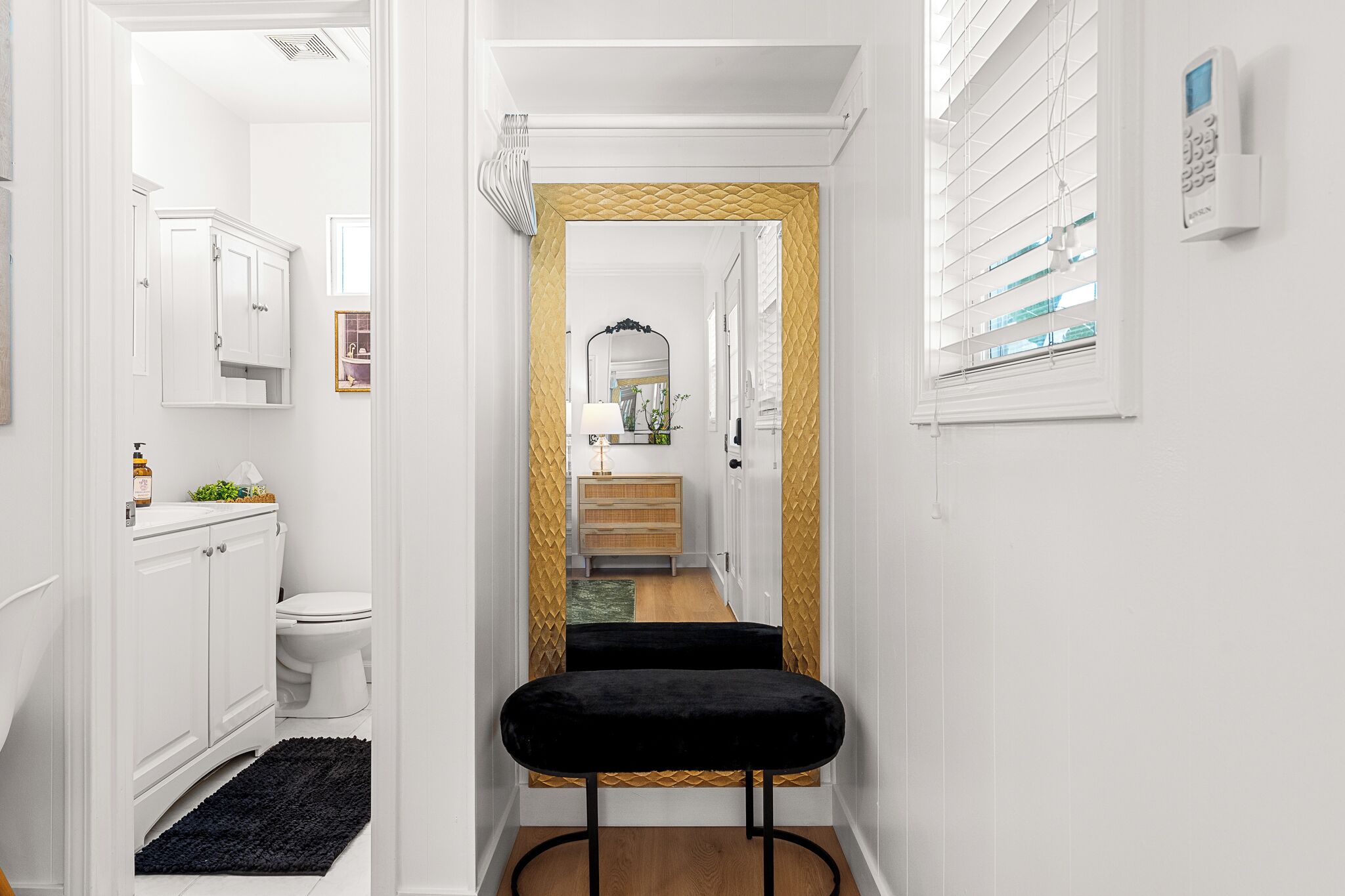 Un couloir étroit avec un banc en velours noir, des meubles blancs, des toilettes et une fenêtre avec des stores.
