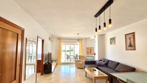 Uma sala de estar moderna com sofá, mesa de centro e televisão. Há uma porta de vidro deslizante que dá para uma área externa.
