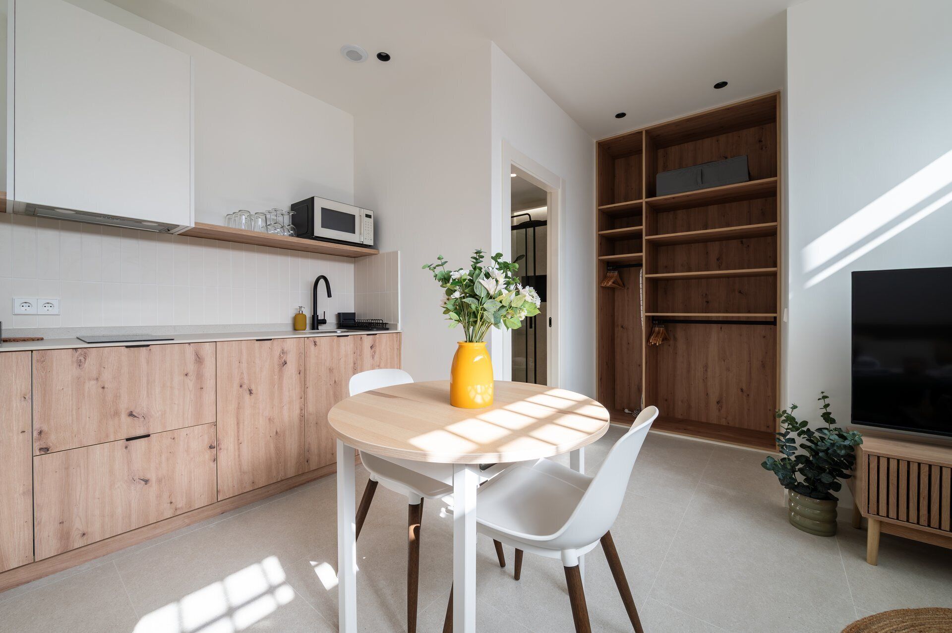 Een moderne keuken met een houten kast, een magnetron en een spoelbak. Er staat een eettafel met stoelen en een vaas met bloemen.