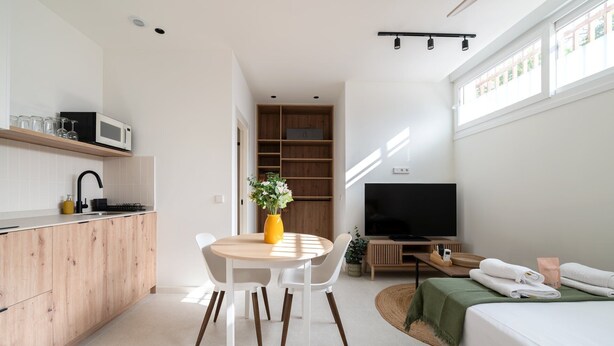 Een ruimtelijk ingerichte leefruimte met kitchenette, eethoek, televisie en bed.