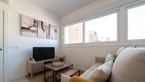 Ein Wohnzimmer mit Fernseher, einem kleinen Tisch, einer Couch und einem Fenster mit Blick auf Gebäude.