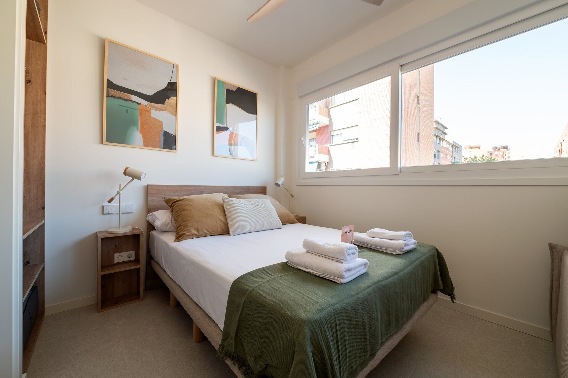 Ein ordentlich eingerichtetes Schlafzimmer mit einem Bett, Nachttischen, einer Lampe, Kunstwerken an der Wand und einem Fenster mit Blick auf Gebäude.