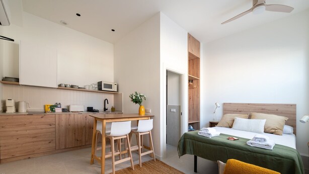 A compact hotel room with a kitchenette, a bed with a green bedspread, a wooden headboard, a small table with two chairs, and a ceiling fan.