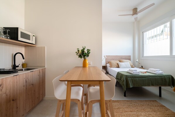A compact room with a kitchenette, a dining table with chairs, a bed with a green bedspread, and a ceiling fan.