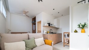 Een moderne woonkamer met een bank, een plafondventilator, een keukenhoek met een magnetron en een kleine tafel met stoelen.