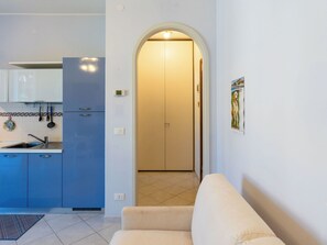 A modern kitchen with blue cabinets, a built-in oven, and a sink. There is a beige sofa in the living area, a door leading to another room, and a tiled floor.