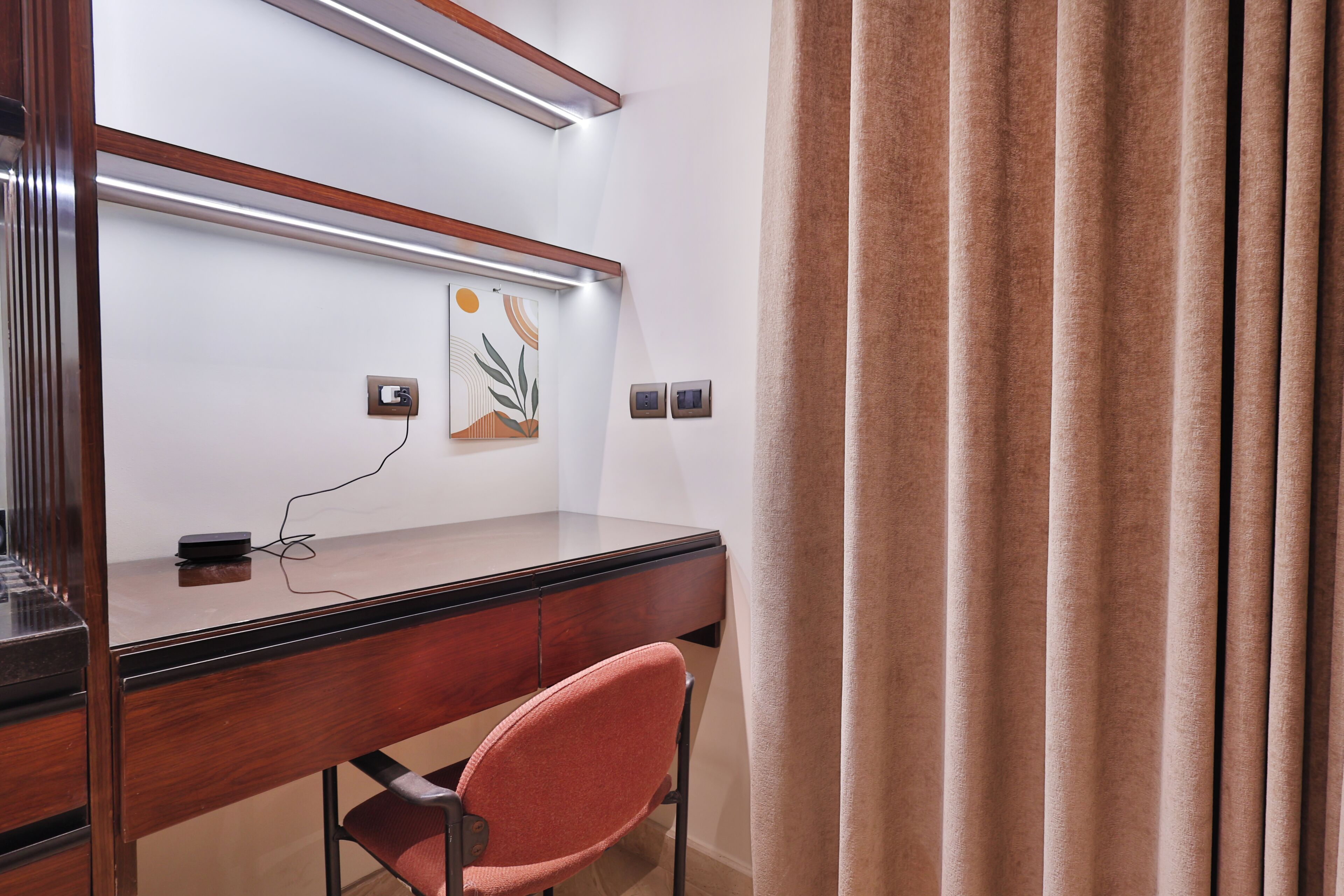 Hotellihuone, jossa on puinen työpöytä, punainen tuoli, seinään kiinnitetty lamppu ja taulu.
