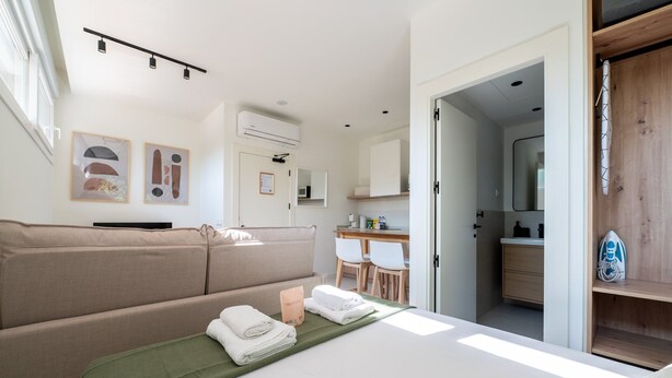 Un dormitorio moderno con cama, colcha verde, sofá, escritorio y baño visible a través de una puerta abierta.
