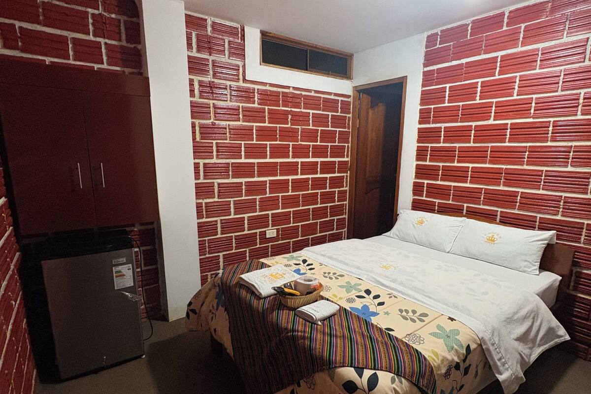 Une chambre avec un mur décoré d’un motif en briques, un lit recouvert d’une courtepointe à motifs floraux, une petite table et un réfrigérateur.