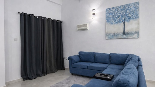 Un salon moderne avec un canapé bleu, un tapis gris, un tableau représentant un arbre et un ventilateur de plafond.