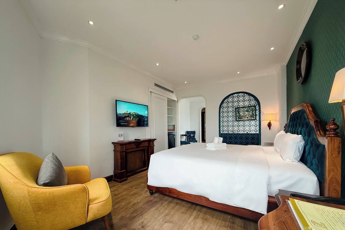 Una camera d'albergo con un letto, una poltrona gialla, una cassettiera in legno, una televisione e un elemento decorativo a parete.