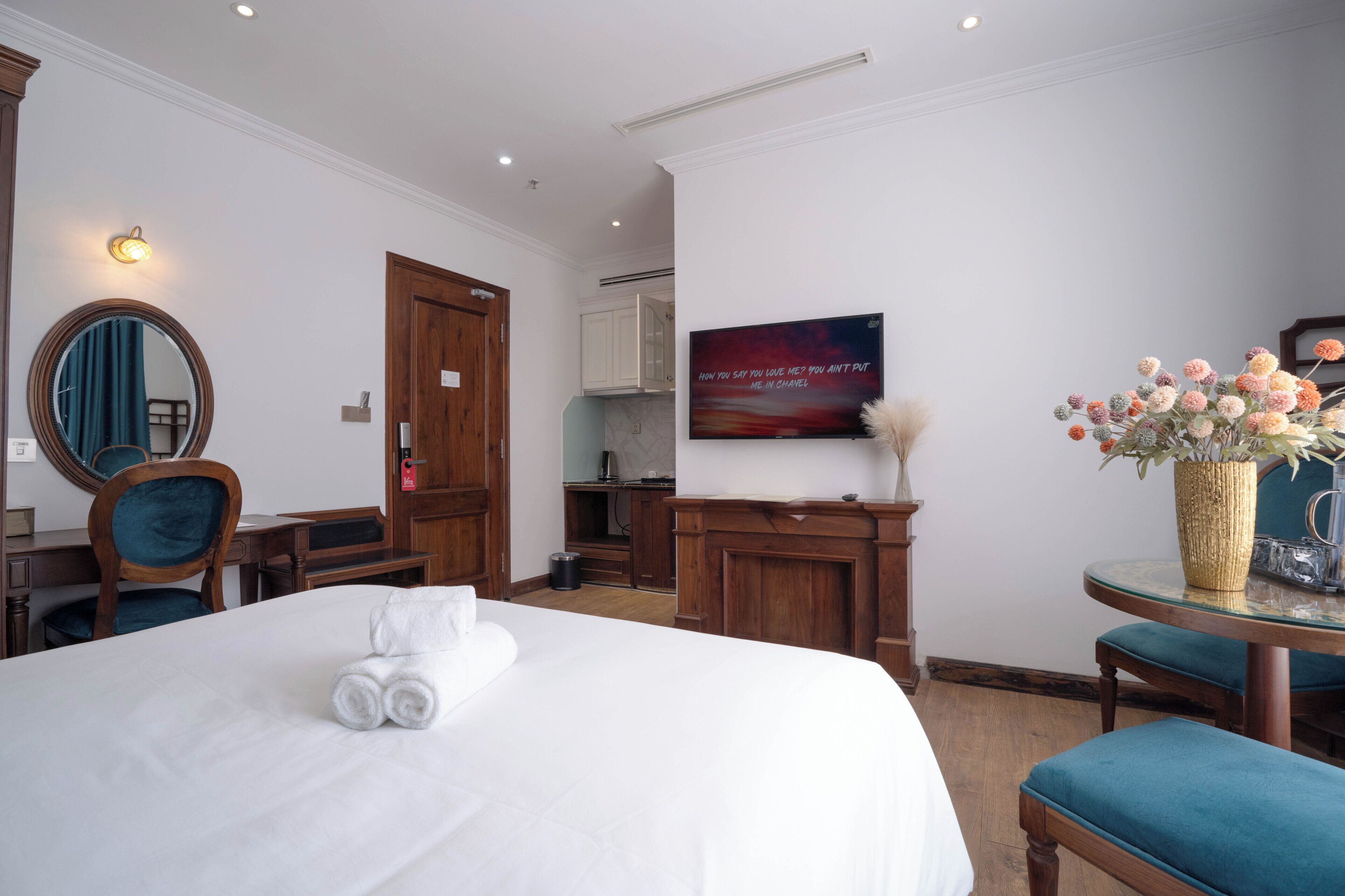 A hotel room with a bed, a desk, a chair, a mirror, a wall-mounted TV, and a vase with flowers.
