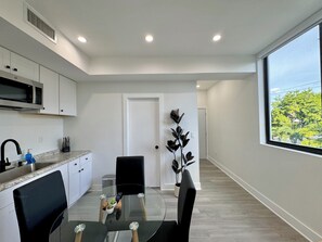 A modern kitchen with white cabinets, a microwave, and a sink. There is a dining area with a glass table and black chairs. A potted plant is on the wall, and a window provides a view of greenery outside. - Havana Studio Suite Near Brickell & Close to Miami Beach (Miami)