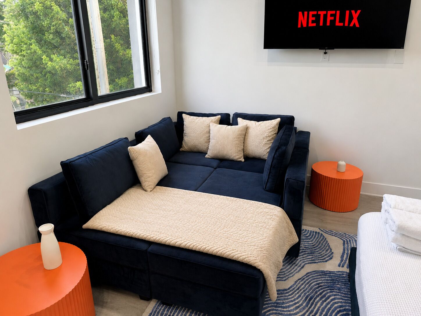 A modern living room with a sofa bed, a TV mounted on the wall, and a window with a view of trees.