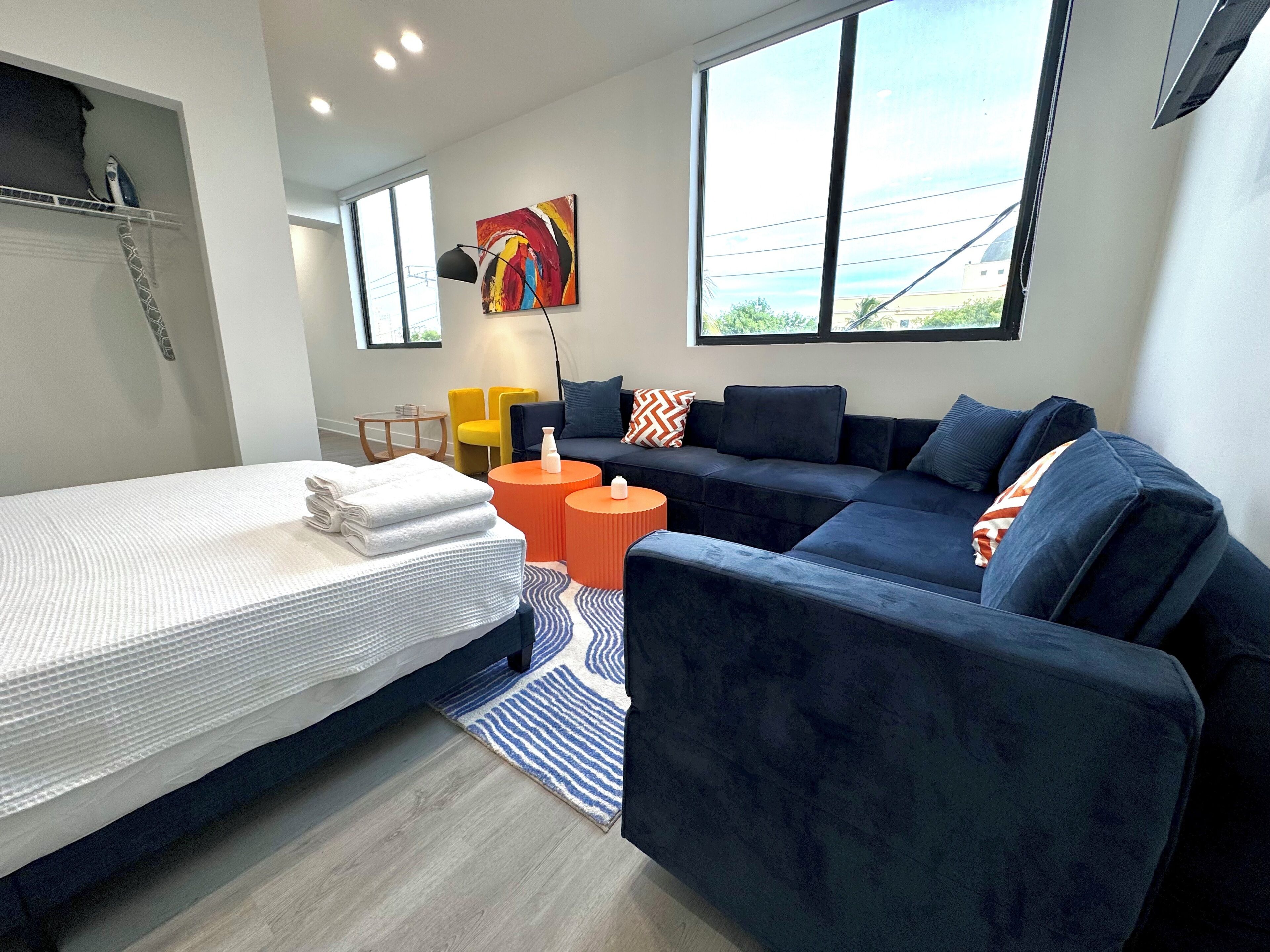 A modern hotel room with a bed, a dark blue sofa, a small orange table, and a colorful abstract painting on the wall.