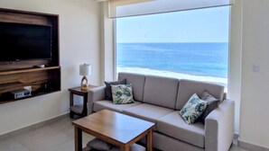 Un salón con sofá, una mesa de centro, un televisor montado en la pared y una ventana con vista al océano.