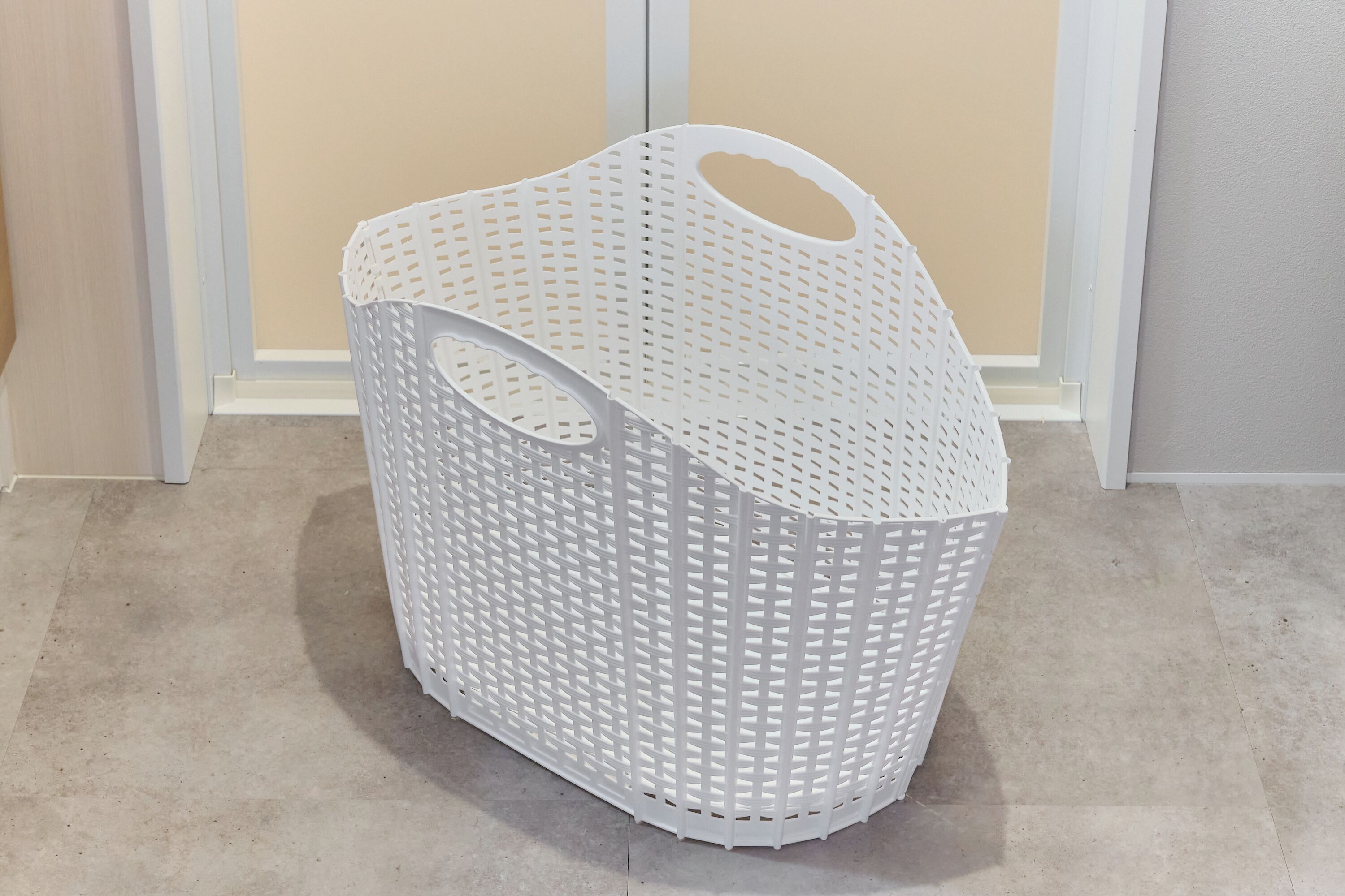 A white plastic laundry basket with a handle, placed on a beige floor.